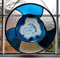 Ten inch round stained glass panel with a blue dyed agate geode center surrounded by alternating turquoise and clear textured cathedral art glass. Chain for hanging is attached.