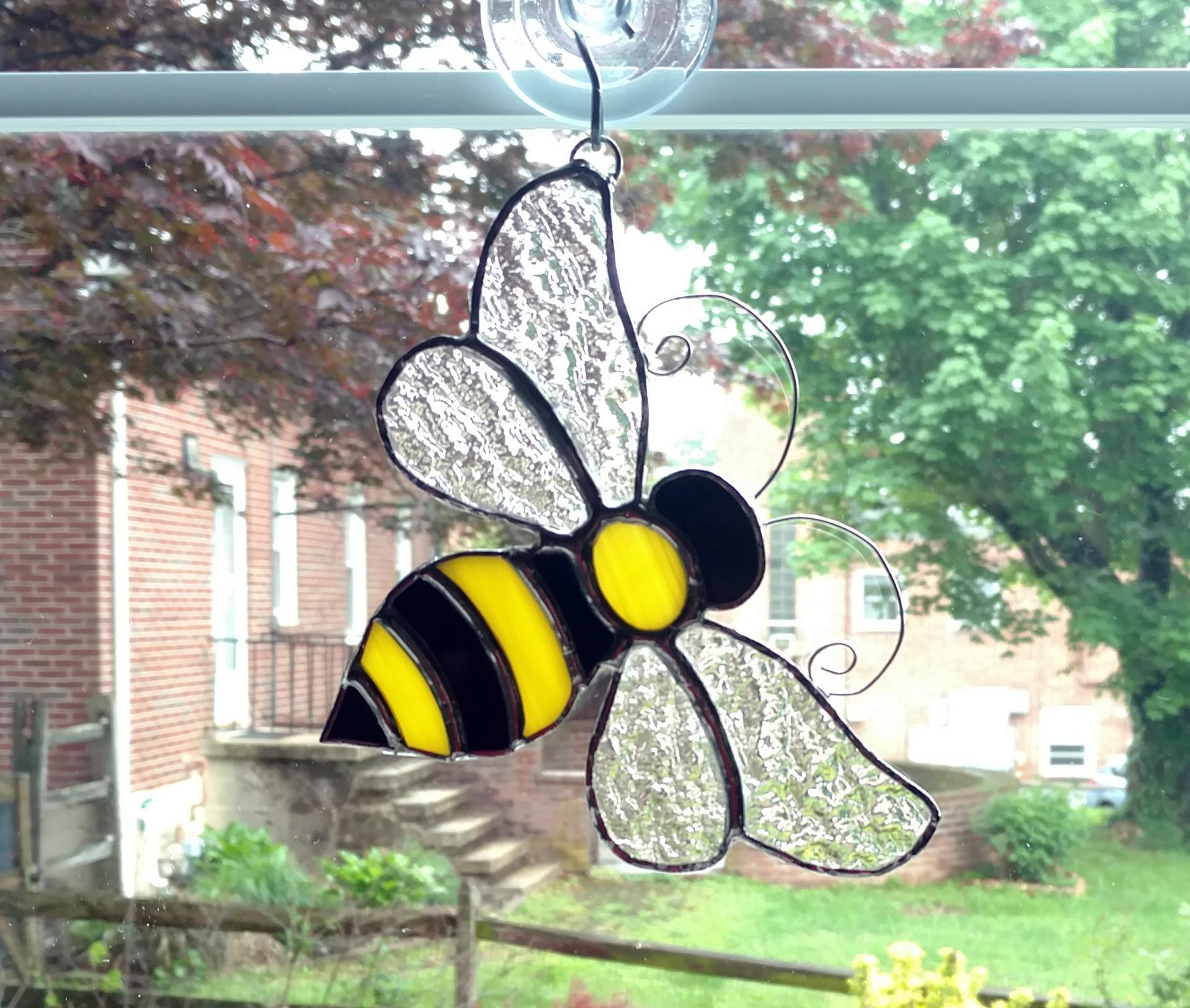 Stained glass bumble bee suncatcher made with black and yellow opalescent art glass for the body and clear textured cathedral glass for the wings.  Curled wire antennae are attached and comes with a suction cup hanger. Measures six inches by five inches.
