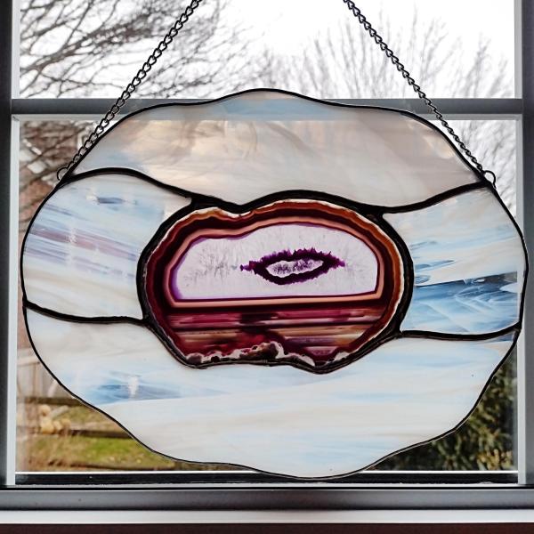Large stained glass suncatcher featuring a purple dyed agate geode in the center that looks like an eye, surrounded by white and clear wispy art glass in a cloud shape. Measures ten inches by eight inches and comes with a chain and suction cup hanger.