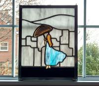 Rectangular stained glass panel depicting a woman facing away in a flowing blue gown holding an umbrella made with a dyed agate geode, overlooking a grey city skyline. Measure nine inches wide by twelve inches tall and is framed in zinc framing with hanging hooks installed in the frame.