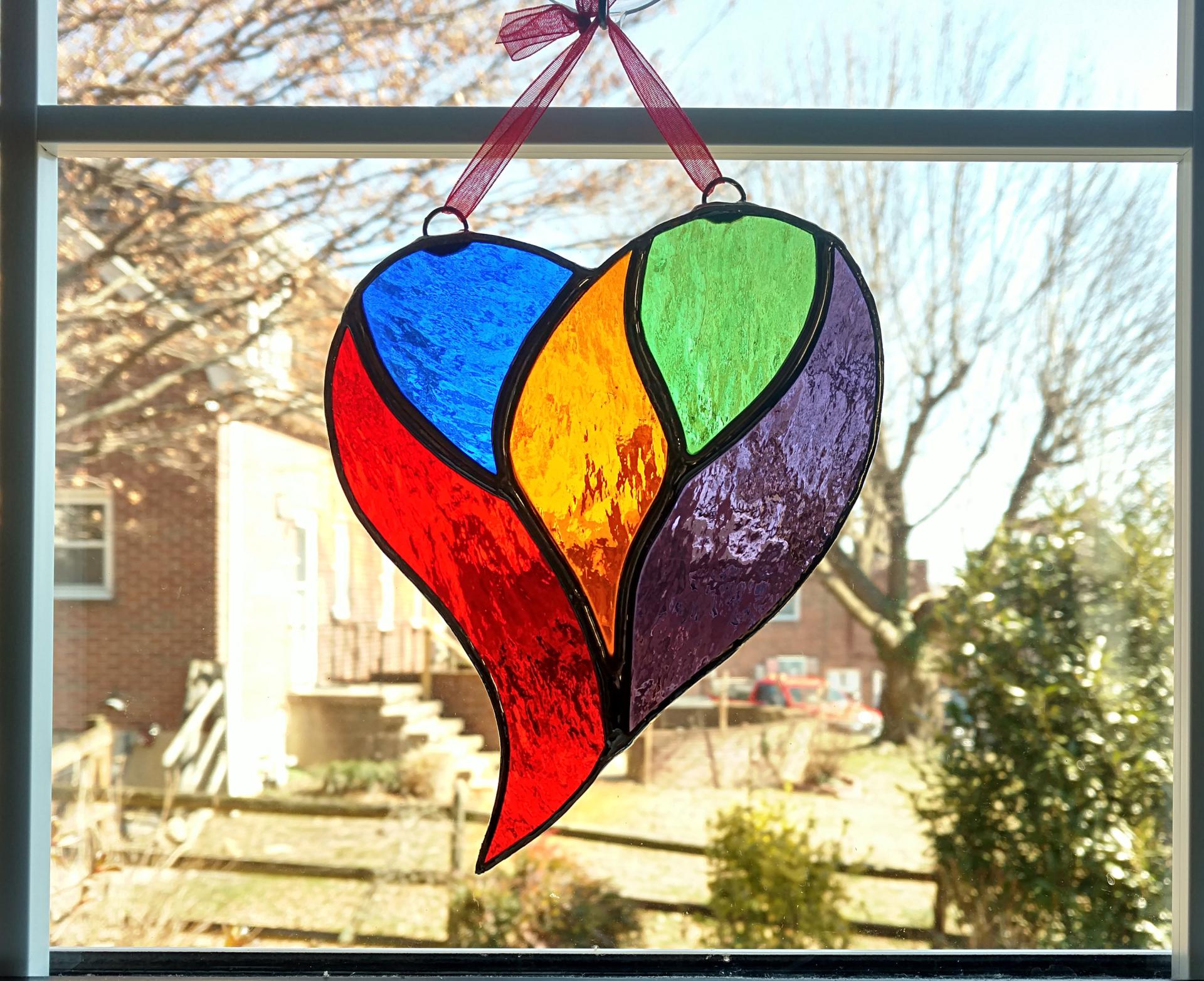 rainbow stained glass heart suncatcher measuring five inches by six inches.  Made with blue, green, amber, purple, and red cathedral glass and comes with a suction cup hanger and red ribbon.