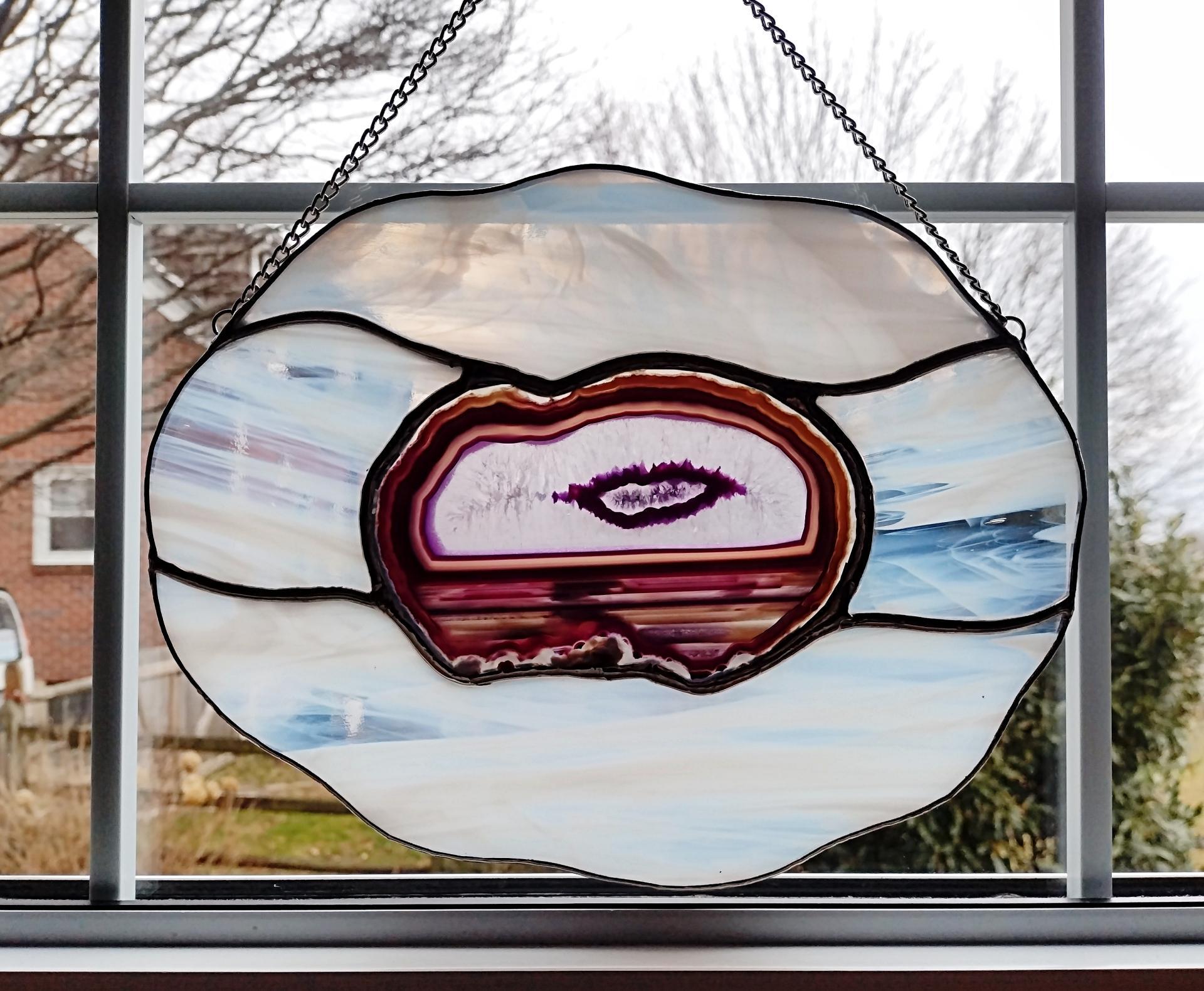 Large stained glass suncatcher featuring a purple dyed agate geode in the center that looks like an eye, surrounded by white and clear wispy art glass in a cloud shape. Measures ten inches by eight inches and comes with a chain and suction cup hanger.