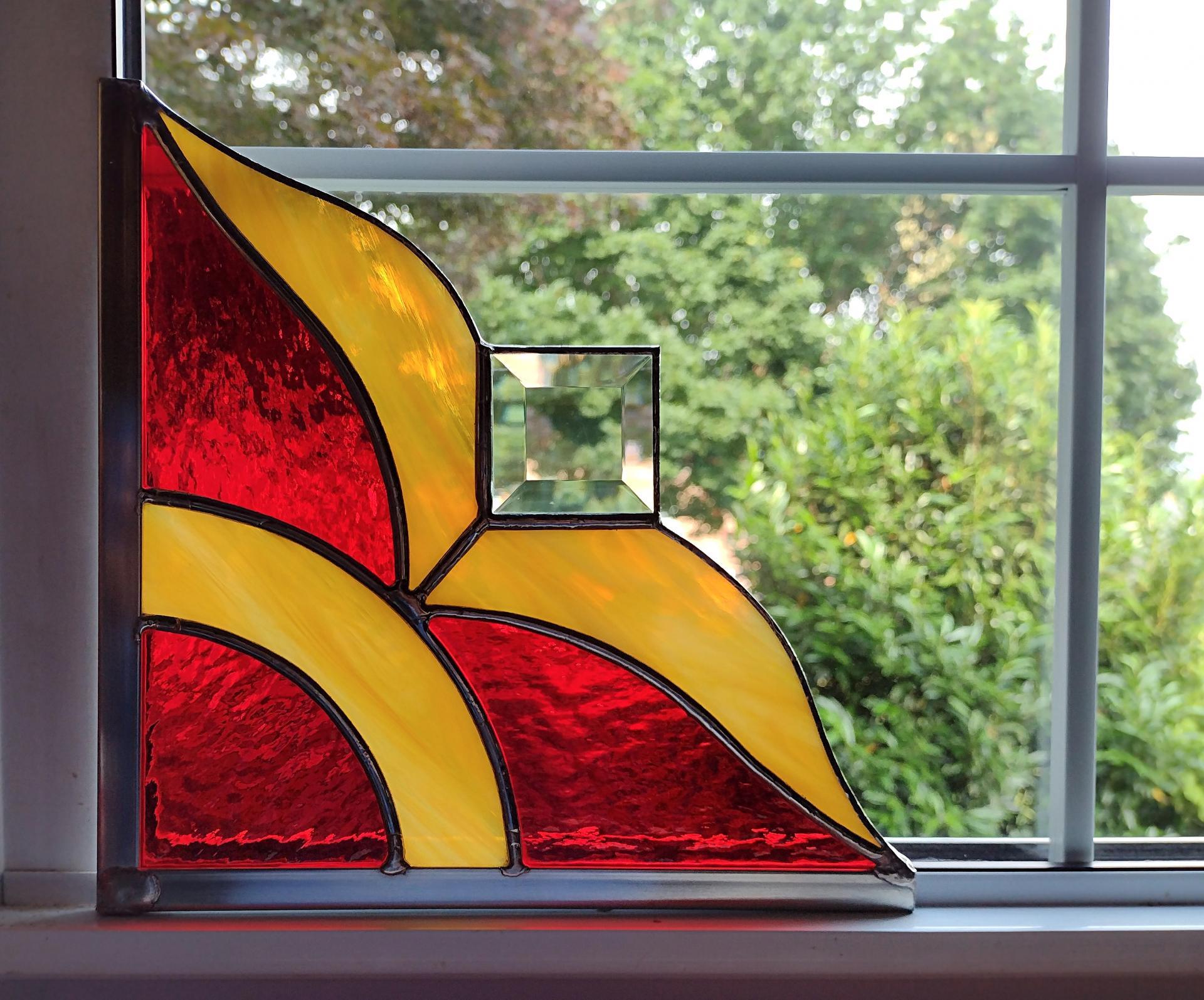 Stained glass corner piece made with alternating red and gold art glass with a clear square bevel prism in the center. Measures nine inches in diameter and is framed on two size in zinc framing for hanging.