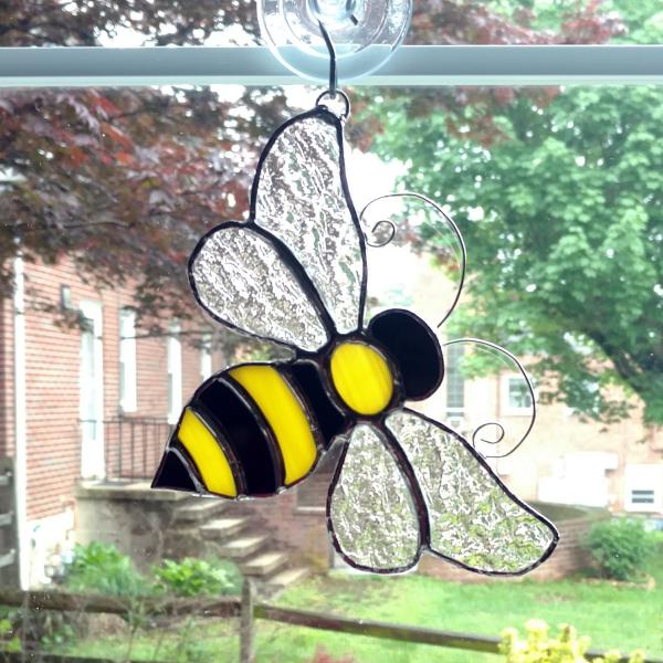 Stained glass bumble bee suncatcher made with black and yellow opalescent art glass for the body and clear textured cathedral glass for the wings.  Curled wire antennae are attached and comes with a suction cup hanger. Measures six inches by five inches.