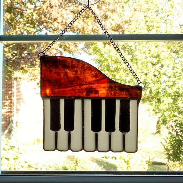 Stained glass piano suncatcher made with a brown swirled opalescent glass top with seven white glass keys and five black glass keys overlaid on top.  Measures six inches wide by five and half inches tall, with a hanging length of eight inches with the attached chain.  Comes with a suction cup hanger.