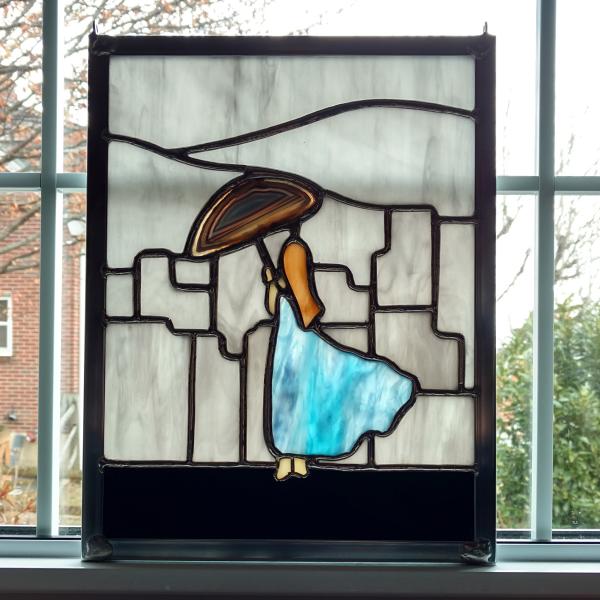 Rectangular stained glass panel depicting a woman facing away in a flowing blue gown holding an umbrella made with a dyed agate geode, overlooking a grey city skyline. Measure nine inches wide by twelve inches tall and is framed in zinc framing with hanging hooks installed in the frame.