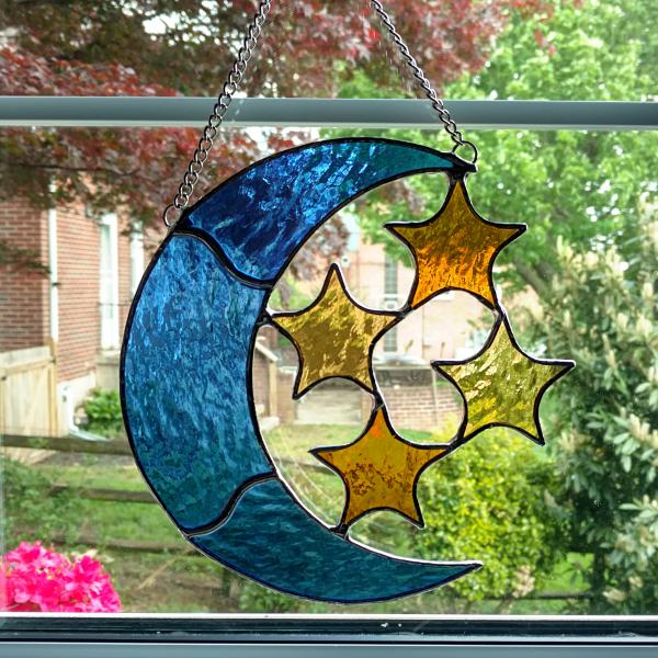 Stained glass blue moon with four amber stars suncatcher made with textured cathedral glass, measuring seven inches by seven inches and comes with chain and suction cup hanger.