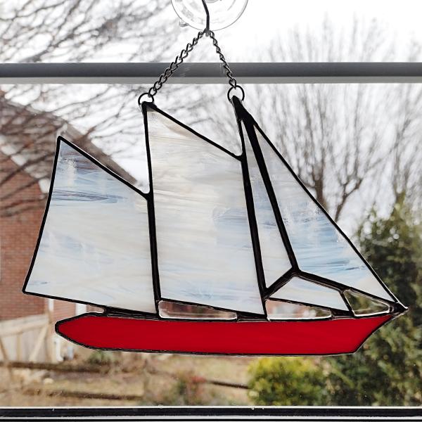 stained glass sailboat schooner suncatcher with a red hull and white and clear swirled sails. 6 in by 8 in plus chain.