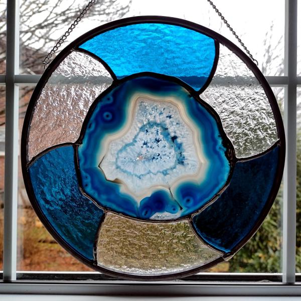 Ten inch round stained glass panel with a blue dyed agate geode center surrounded by alternating turquoise and clear textured cathedral art glass. Chain for hanging is attached.