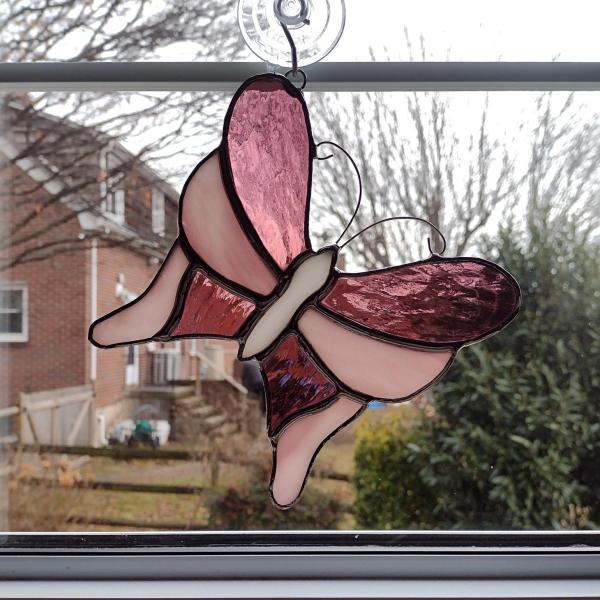 Pink stained glass butterfly suncatcher, made with alternating pink cathedral glass and pink opalescent glass with a white glass body. It measures five and half inches in diameter and comes with a suction cup hanger.