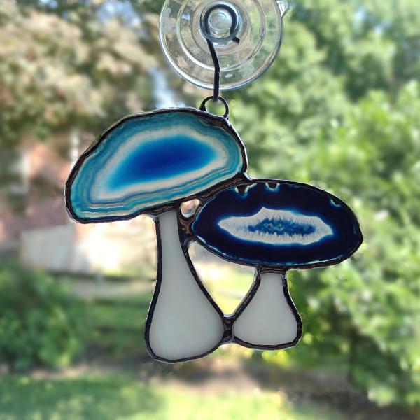 Group of two mushrooms suncatcher with mushroom caps made from blue dyed agate geodes and stems made with white opalescent art glass. Measure three inch by four inches and comes with a suction cup hanger.