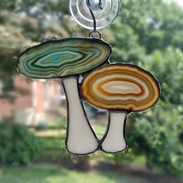 Agate geode mushroom cluster suncatcher with blue and brown dyed agate geodes for the caps and white opalescent glass for the stems. Measures four inches by five inches and comes with a suction cup hanger.