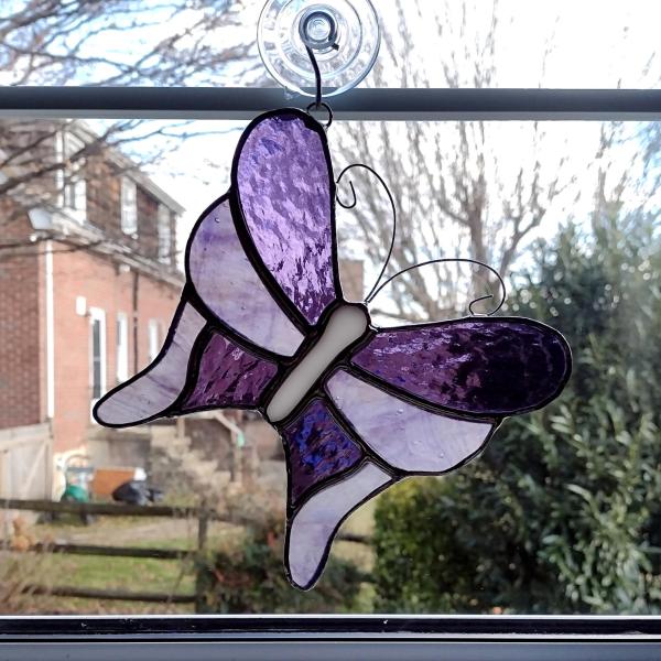 Stained glass butterfly suncatcher made with alternating purple cathedral glass and purple swirled opalescent glass with curled wire antennae.  Measures five and half inches in diameter and comes with a suction cup hanger.