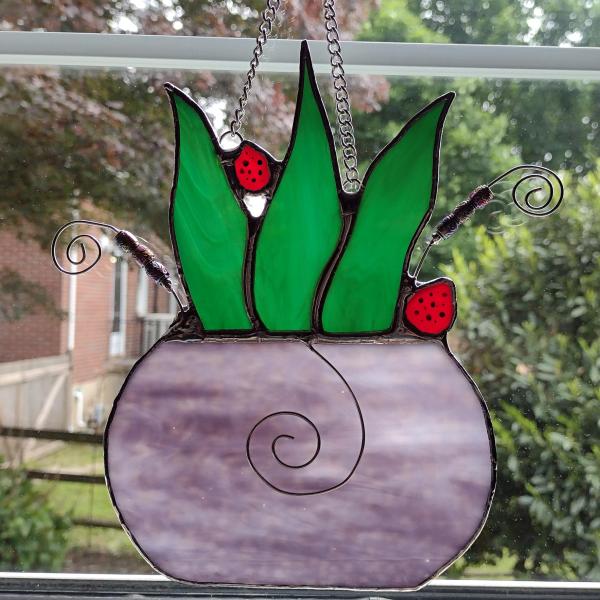 stained glass potted snake plant with two ladybugs perched, measures eight inches by seven inches, chain and suction cup hanger included.