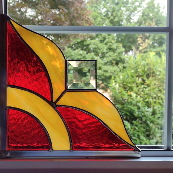 Stained glass corner piece made with alternating red and gold art glass with a clear square bevel prism in the center. Measures nine inches in diameter and is framed on two size in zinc framing for hanging.