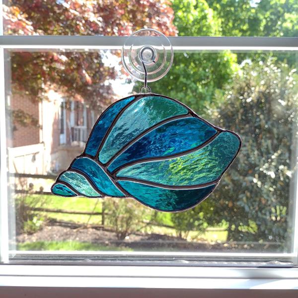 Stained glass conch shell suncatcher made with alternating turquoise and light aqua textured cathedral art glass. Measures seven inches wide by four and half inches tall and comes with a suction cup hanger. 