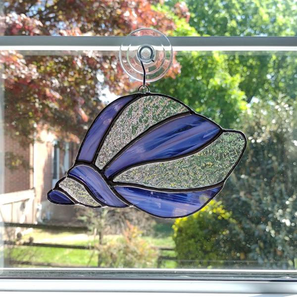 Stained glass conch shell suncatcher made with alternating purple and white swirled art glass and clear textured cathedral art glass. Measures seven inches by four and half inches and come with a suction cup hanger.