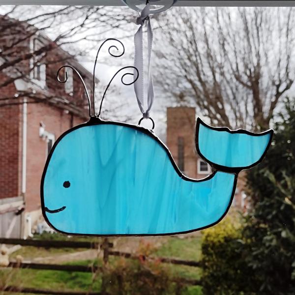 Stained glass blue whale suncatcher measuring five and half inches diameter, with curled wire water spouting. Suction cup hanger included.