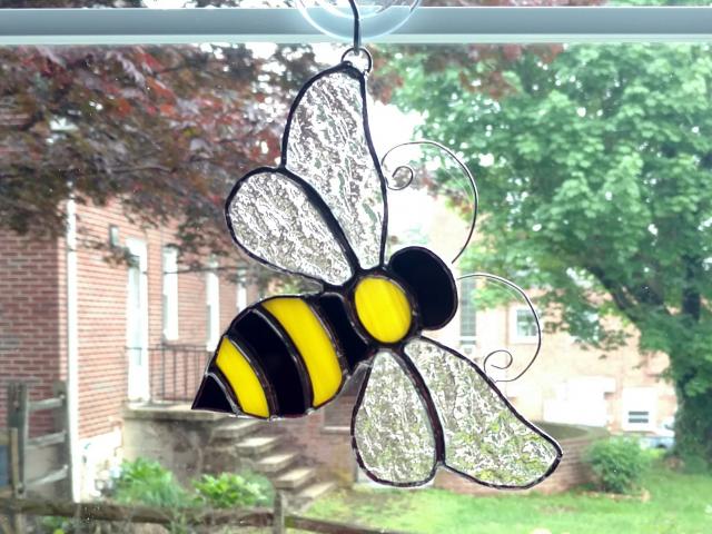 Stained glass bumble bee suncatcher made with black and yellow opalescent art glass for the body and clear textured cathedral glass for the wings.  Curled wire antennae are attached and comes with a suction cup hanger. Measures six inches by five inches.