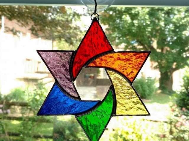 Stained glass star of David made with rainbow colors of red, orange, yellow, green, purple, and blue textured cathedral art glass.  Measures six inches in diameter and comes with a suction cup hanger.