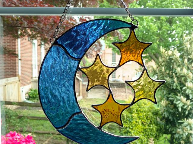 Stained glass blue moon with four amber stars suncatcher made with textured cathedral glass, measuring seven inches by seven inches and comes with chain and suction cup hanger.