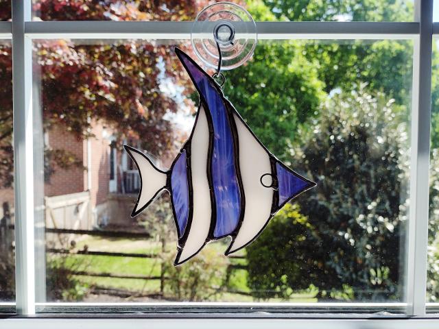 Stained glass angel fish suncatcher made with alternating purple and white wispy glass and white iridescent glass. Suction cup hanger included. Measures five inches by 6 inches.