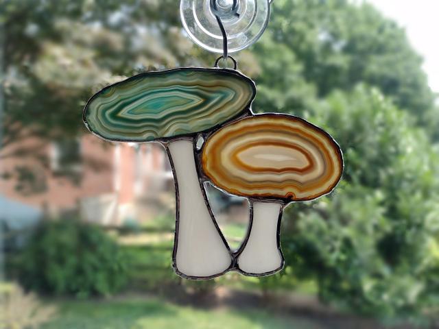 Agate geode mushroom cluster suncatcher with blue and brown dyed agate geodes for the caps and white opalescent glass for the stems. Measures four inches by five inches and comes with a suction cup hanger.