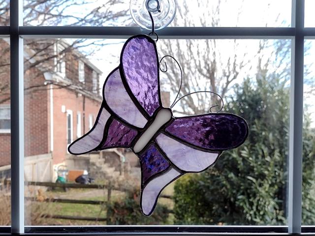 Stained glass butterfly suncatcher made with alternating purple cathedral glass and purple swirled opalescent glass with curled wire antennae.  Measures five and half inches in diameter and comes with a suction cup hanger.