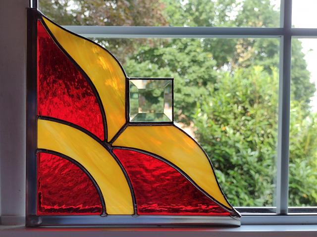 Stained glass corner piece made with alternating red and gold art glass with a clear square bevel prism in the center. Measures nine inches in diameter and is framed on two size in zinc framing for hanging.