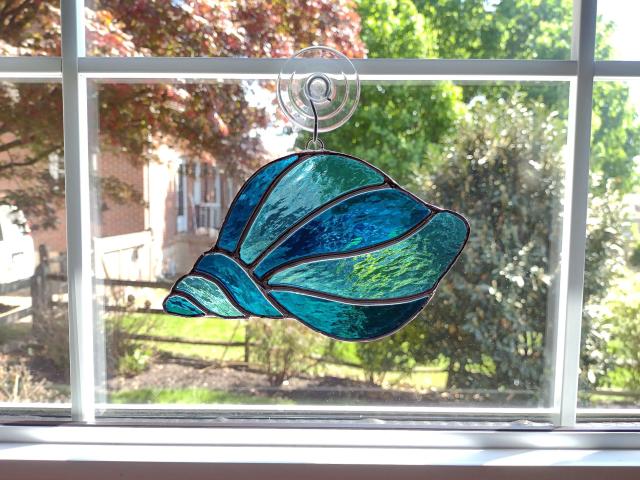 Stained glass conch shell suncatcher made with alternating turquoise and light aqua textured cathedral art glass. Measures seven inches wide by four and half inches tall and comes with a suction cup hanger. 