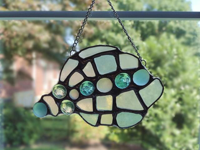 conch shell suncatcher made with pale blue, green, and yellow sea glass and glass jewels, measures approximately seven inches by four inches, attached chain and suction cup holder included.